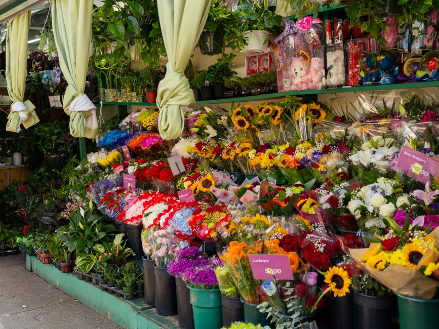 A vibrant flower display outside a florist shop, featuring an assortment of fresh flowers including sunflowers, roses, lilies, and mixed bouquets wrapped in colorful paper and cellophane. The flowers 