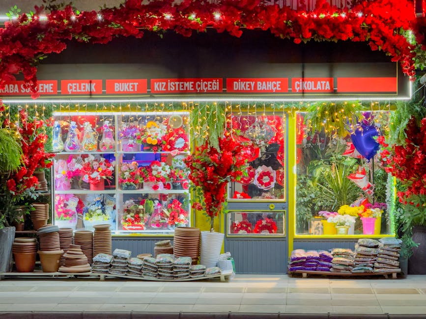 A vibrant floral shop exterior featuring a brightly lit display window filled with various colorful flower arrangements, including roses, tulips, and carnations in shades of red, pink, purple, and whi