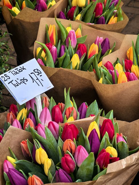 Multiple bouquets of fresh tulips in vibrant shades of pink, yellow, purple, red, and orange, wrapped in brown craft paper, are arranged on a wooden surface. A handwritten sign indicating the delivery