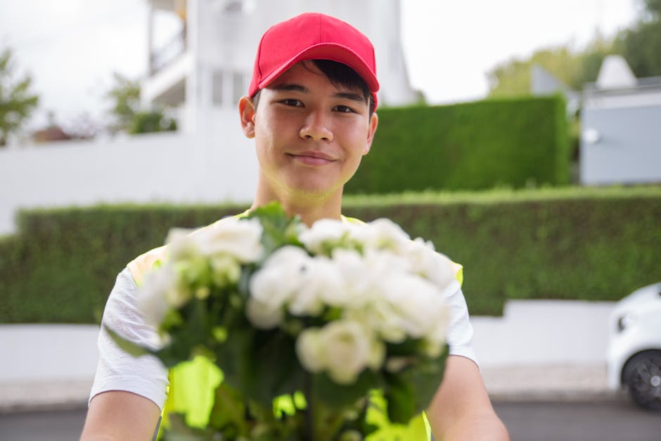 A delivery person wearing a red cap and a high-visibility yellow vest is standing outdoors, holding a bouquet of fresh white flowers with green foliage. The bouquet is prominently displayed in the for