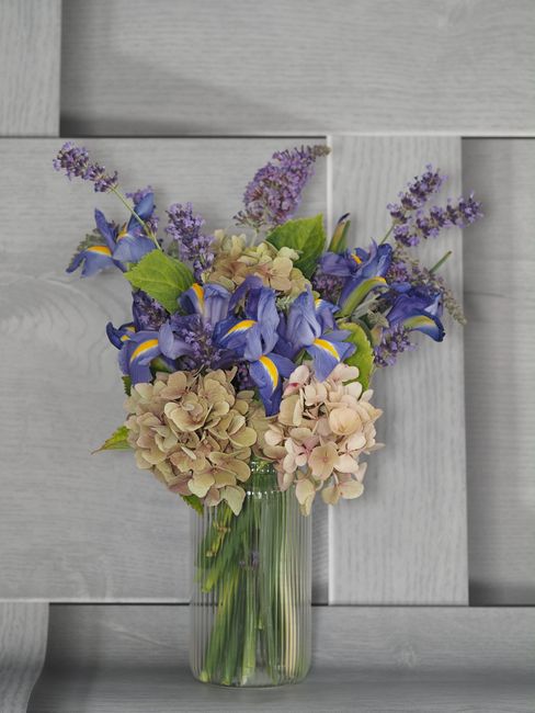 A tall, clear glass vase filled with a floral arrangement featuring cream-colored hydrangeas, purple irises, and sprigs of lavender. The bouquet is set against a light grey wooden background, with the