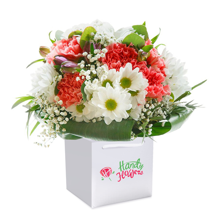 Set atop a seamless white stage, this round, hand-tied bouquet blossoms from within a spotless square gift bag-a testament to Friern Barnet's discerning floral artistry. A medley of coral, peach, and ruby red carnations stretch upward, their lacy, ruffled petals reflecting the gentle transition from dusk to dawn across the greenery of North London. The arrangement is punctuated by crisp, white daisies, each blossom crowned with a zesty lime-green center, weaving a thread of innocence and warmth throughout. Baby's breath is generously tucked between the more prominent flowers, its frothy white sprays lending a textural softness, like morning mist over Bethune Park. Around the circumference, broad, gleaming green leaves unfurl in elegant arcs, forming a natural, tailored collar that gives substance and coherence to the design. Look closely and you'll spot slender, closed buds blushed with a hint of purple-perhaps alstroemeria-adding understated complexity and keeping the bouquet intriguing long after the first glance. The white gift bag, smartly designed with robust ribbon handles, holds the arrangement upright and secure, symbolizing readiness for delivery from Friern Barnet Flowers-the trusted local source for timely, fresh floral gifts. This image distills the florist's devotion to both form and freshness, making it a perfect visual for articles celebrating gift-giving occasions, neighbourhood traditions, or the convenience of flower delivery woven through Friern Barnet's daily life.