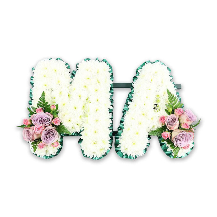 This image presents a dignified funeral letter tribute spelling out the word MA, thoughtfully designed as a personal message of remembrance. Set against a plain white backdrop, the letters stand out with crisp clarity, their forms solid and reassuring. Each stroke of the M and the A is densely packed with pristine white chrysanthemums; the blossoms are small but abundant, their layered petals creating a velvety texture that suggests softness and care. No stem or base shows through, just an uninterrupted expanse of white, symbolising purity, peace, and a gentle farewell. Around the perimeter of each letter runs a deep emerald green ribbon, expertly arranged into a slight ruffle that hugs every corner and angle. This border gives definition and contrast, neatly separating the white flowers from the surrounding space and lending the design a refined, finished look. At the lower left base of the M and the lower right base of the A, two matching arrangements add a note of colour and tenderness. These are composed of full-headed pink roses, muted lilac roses, and smaller pink spray roses in various stages of bloom, from tight bud to open rosette. Light, arching fern fronds emerge from among the petals, their fine texture echoing the greenery one might see in the gardens and parks around Friern Barnet. The tribute has a calm, contemplative presence, clearly intended for a funeral or memorial service where a mother, mama, or maternal figure is being honoured. It conveys both respect and intimacy, like a final whispered thank you crafted in flowers.