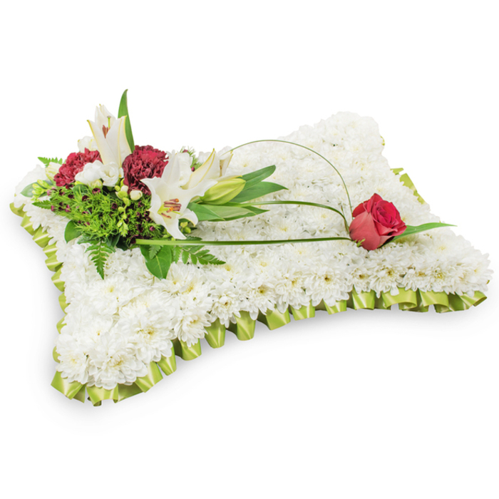 Presented on a clean white surface, this rectangular floral cushion is a traditional and tender tribute shaped with careful florist's skill. The whole body of the pillow is formed from thousands of small white chrysanthemum blooms, packed to create a uniform, velvet-like plane that reads as serenity and rest. Surrounding the perimeter, a neat pleated band of pale lime green ribbon (echoing the fresh green of early spring) frames the composition with a soft, refined border. On the upper left a layered cluster of accent flowers rises naturally: two open white Oriental lilies showing their yellow stamens, a closed lily bud adding a vertical note, and groups of deep burgundy spray carnations that bring warmth and textural contrast. Foliage in varying greens - ferny, glossy and matte leaves - provides depth and a sense of shelter behind the blooms. From the left cluster a graceful arc of green salal or slender grass is drawn across the chrysanthemum bed, forming an elegant line that culminates in a single deep red rosebud resting on the upper right corner of the pillow; the rosebud's velvety sheen and tight petals suggest abiding love and respectful remembrance. The lighting is soft and neutral, highlighting petals' contours while keeping the overall mood composed and reverent. This sympathy cushion, suitable for funeral chapels, crematorium services or private wakes in Friern Barnet and nearby streets such as Friern Barnet Road and Colney Hatch Lane, combines classic flowers and careful design to convey heartfelt condolence.